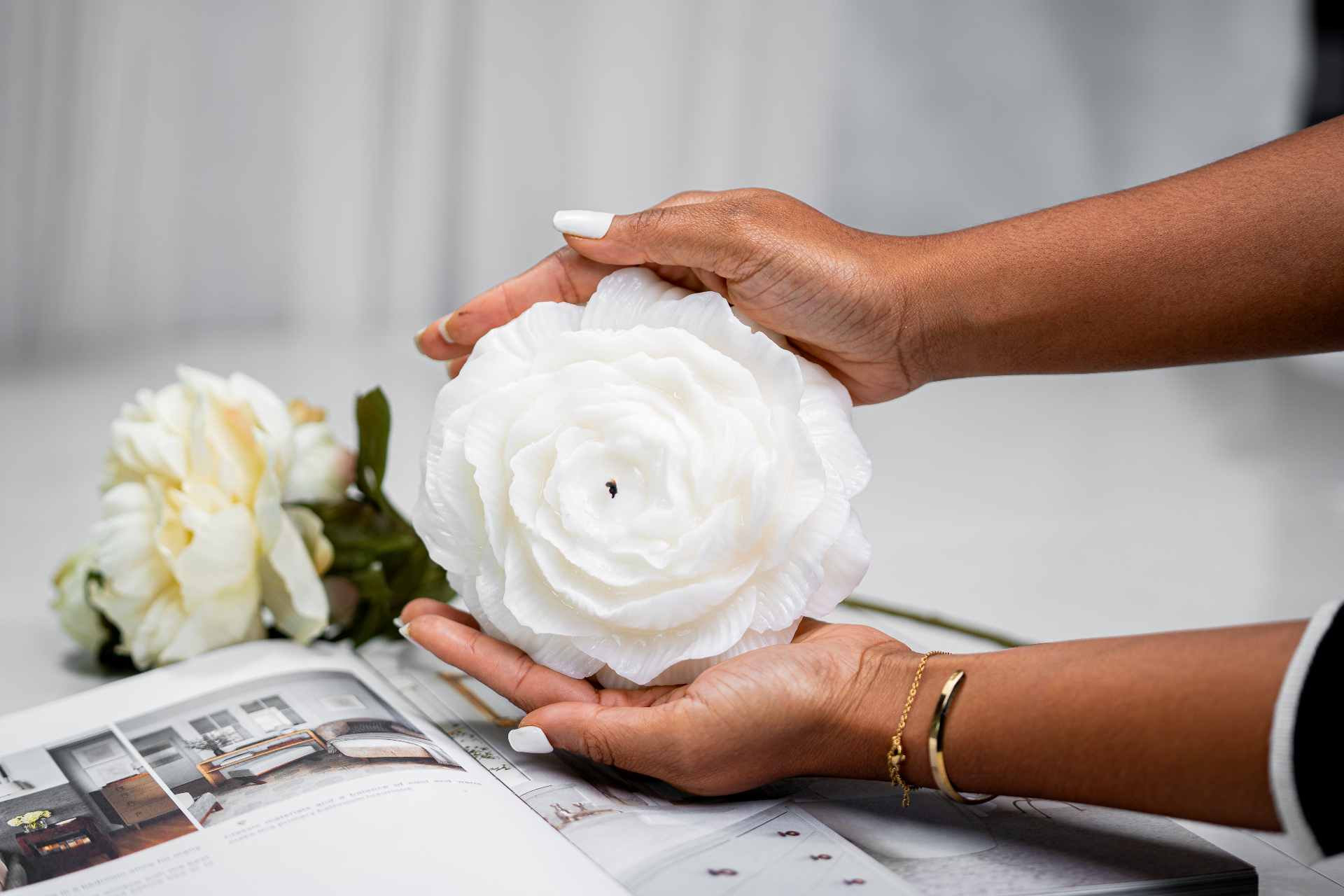 Hands gently holding a white rose LargeBloom candle over an open magazine, with soft peonies nearby